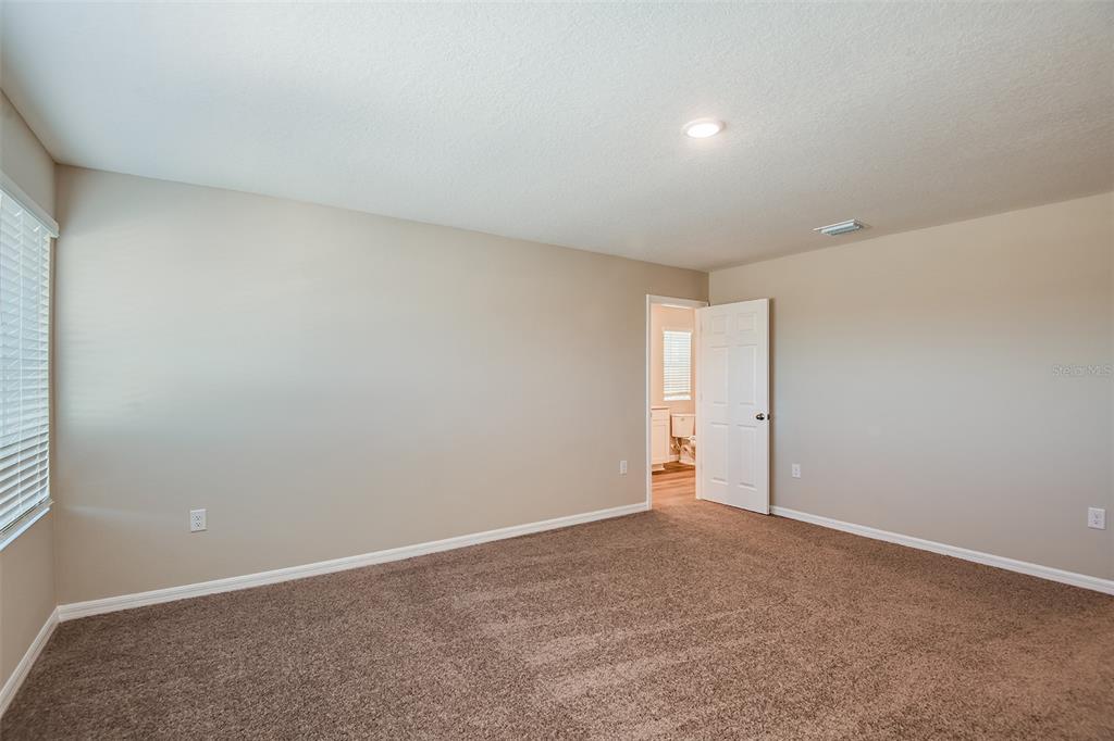 A spacious bedroom with beige walls, brown carpeting, and an open door to a bathroom — 5437 SAN PIETRO DRIVE, WINTER HAVEN, FL