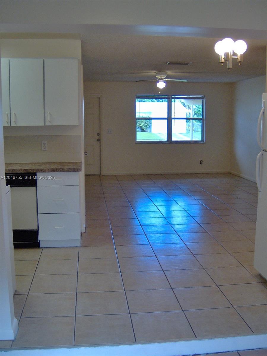 A kitchen with white cabinets and tile flooring opens into a living area with windows — PALM BEACH GARDENS, FL