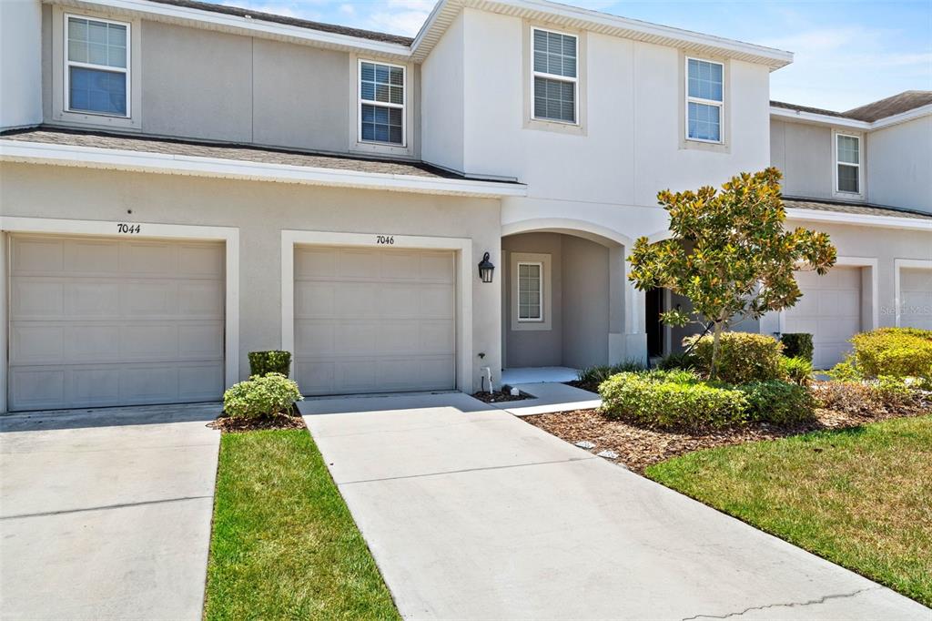 A two-story townhouse exterior features two garage doors, light-colored siding, and a small front yard — 7046 GRAND ELM DRIVE, RIVERVIEW, FL