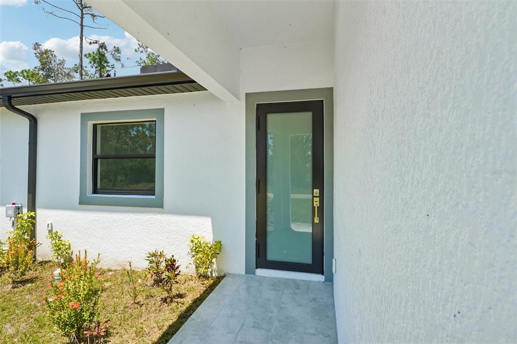 A modern glass-paneled front door sits on a tiled porch next to a white wall — 845/847 MEADOW ROAD, LEHIGH ACRES, FL