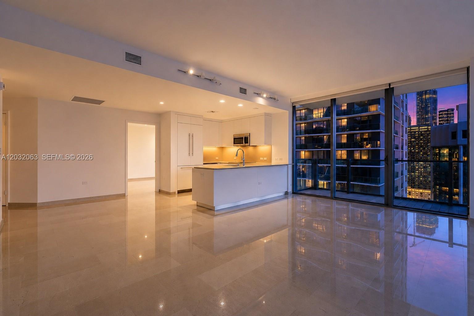A modern kitchen with white cabinetry and an island overlooks a city skyline at dusk — 88 SW 7th St # 2302, Miami FL 33130, MIAMI, FL