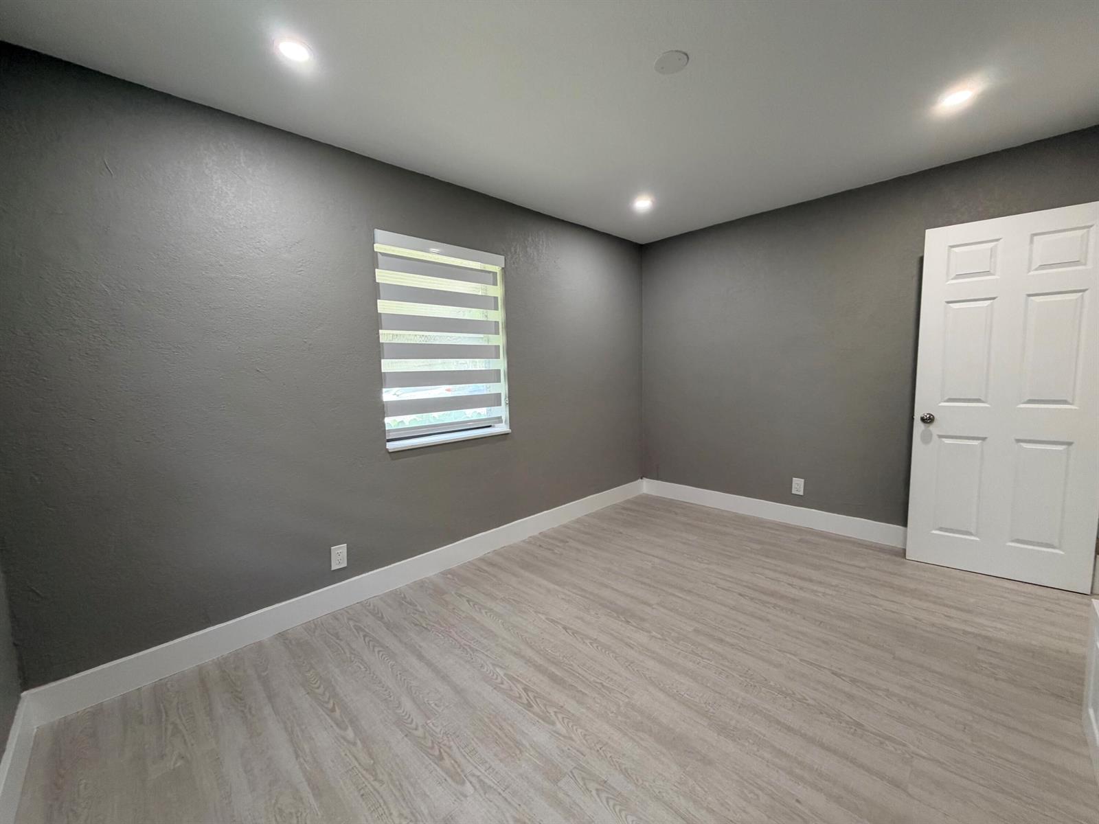 An empty room features gray walls, light wood-look flooring, a window, and a white door — 827 Franklin Road, West Palm Beach FL 33405, WEST PALM BEACH, FL