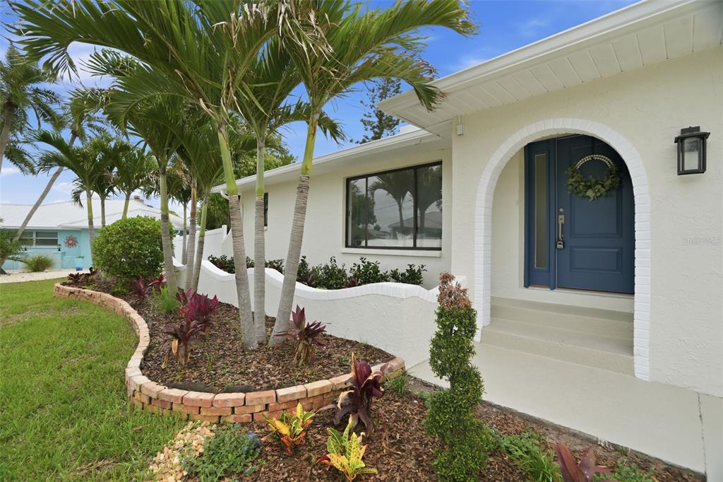 A white house exterior features a blue front door, arched entryway, and a palm-filled garden — 604 KINGSTON COURT, APOLLO BEACH, FL