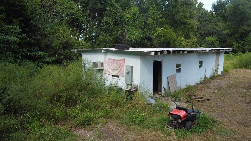 A weathered, single-story white structure sits in a grassy, wooded lot with a riding mower — 17281 SE 130TH, WEIRSDALE, FL