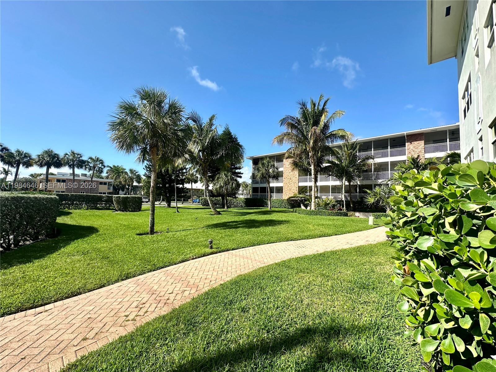 A paved walkway winds through a grassy courtyard with palm trees and apartment buildings — 2501 S Ocean Blvd # 1040, Boca Raton FL 33432, BOCA RATON, FL
