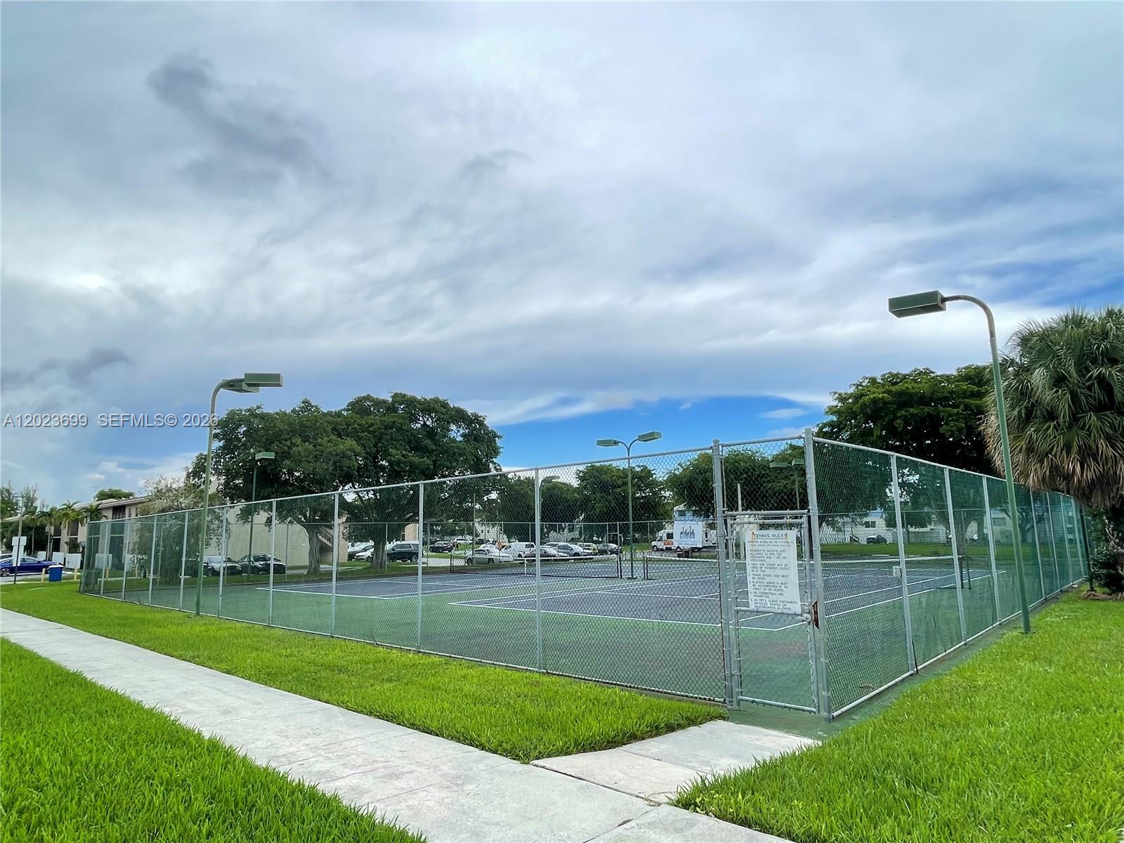 A fenced outdoor tennis court is surrounded by grass and trees under a cloudy sky — 10370 SW 220th St # 224, Cutler Bay FL 33190, CUTLER BAY, FL