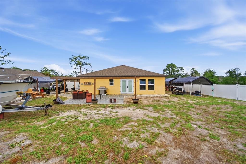 The backyard of a yellow house features a stone patio, a wooden pergola, and carports — 10198 ELGIN BOULEVARD, SPRING HILL, FL