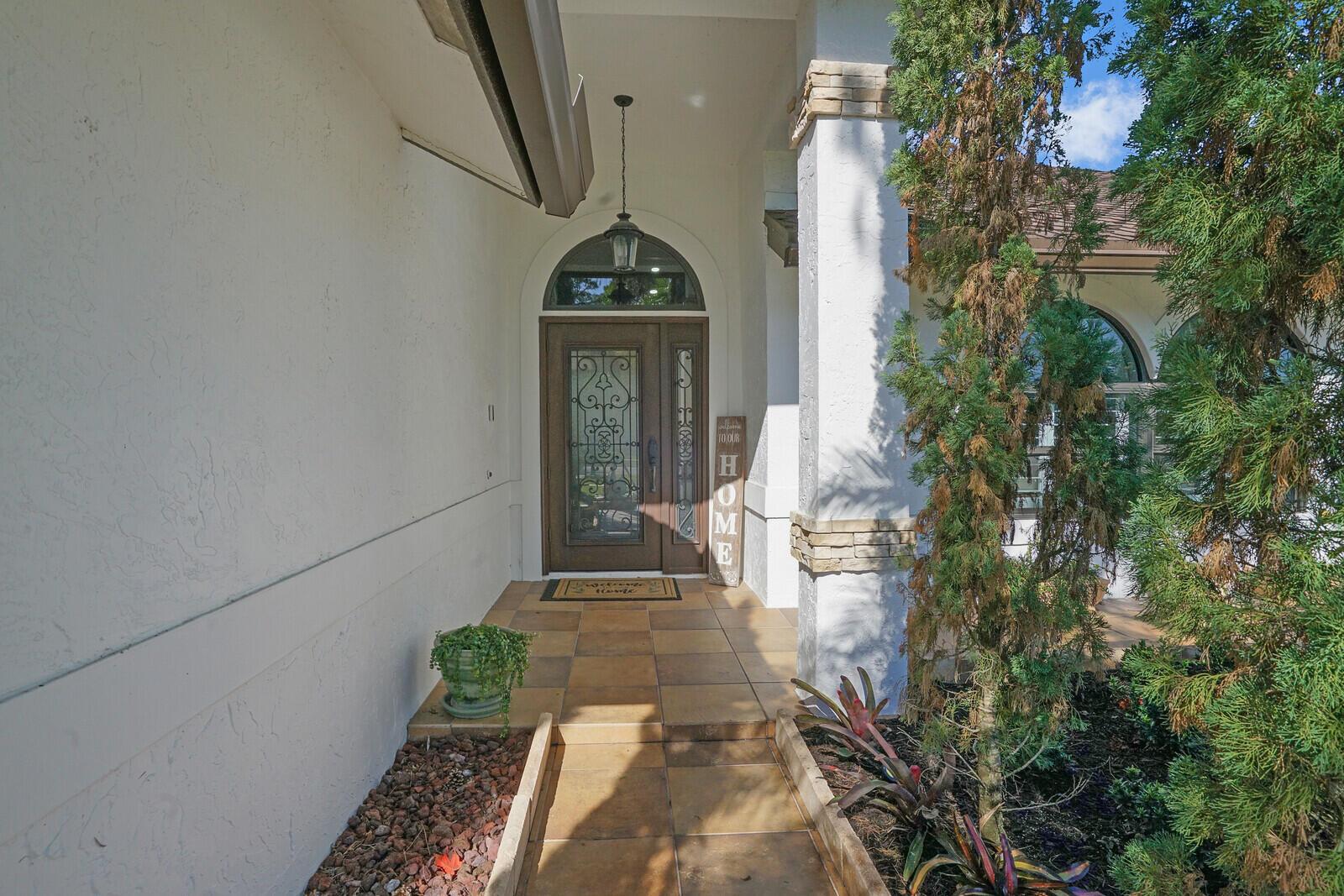 A covered entryway features a tiled walkway, a decorative glass front door, and stone columns — 1313 Brampton Cove, Wellington FL 33414, WELLINGTON, FL