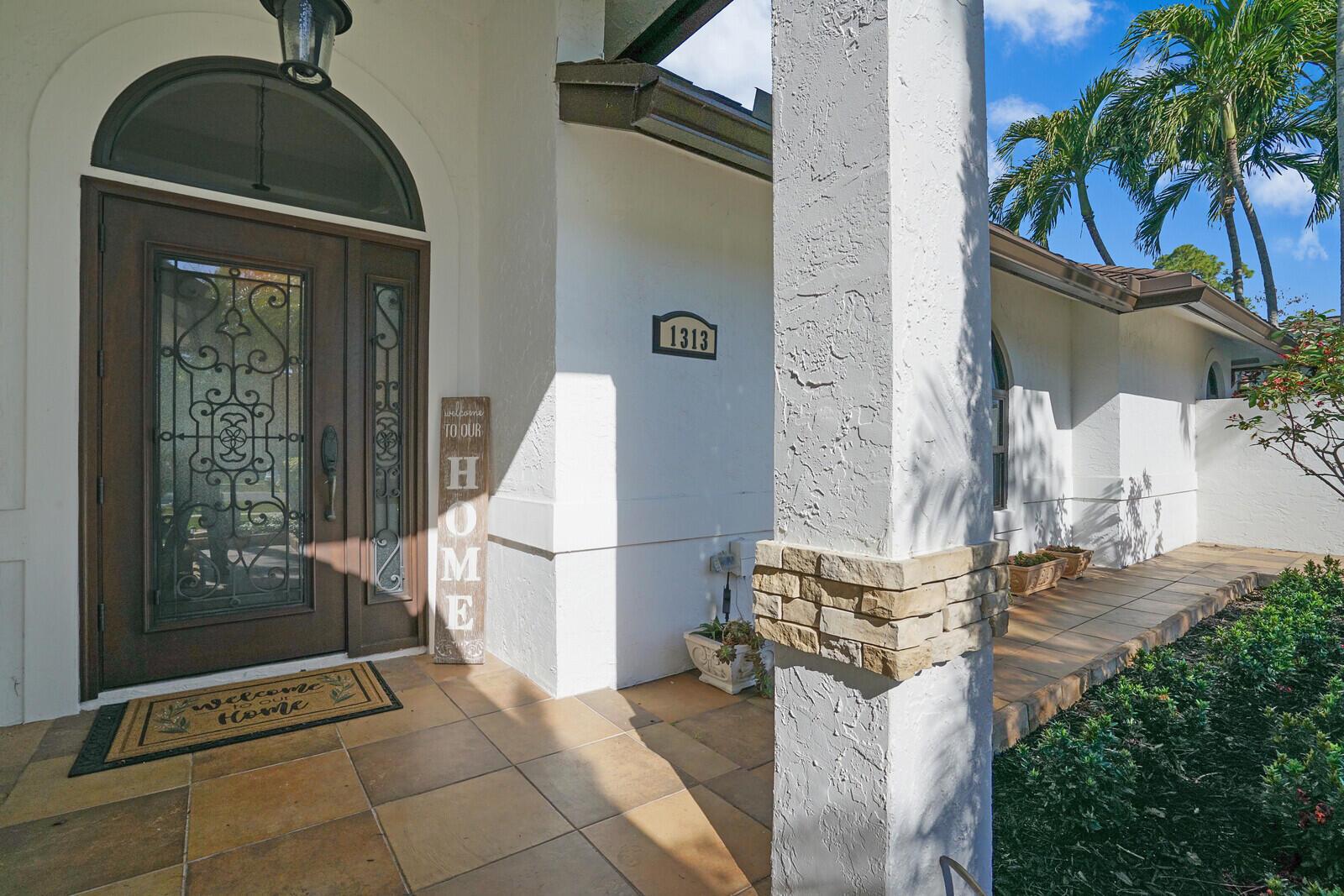 A tiled entryway features a dark wood front door with decorative ironwork and a column — 1313 Brampton Cove, Wellington FL 33414, WELLINGTON, FL