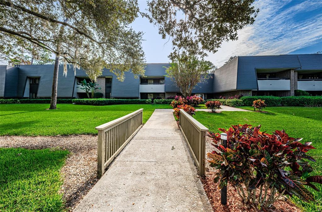 A concrete footbridge leads toward the entrance of a multi-story apartment building with landscaping — 36750 US HIGHWAY 19 N UNIT 5-104, PALM HARBOR, FL