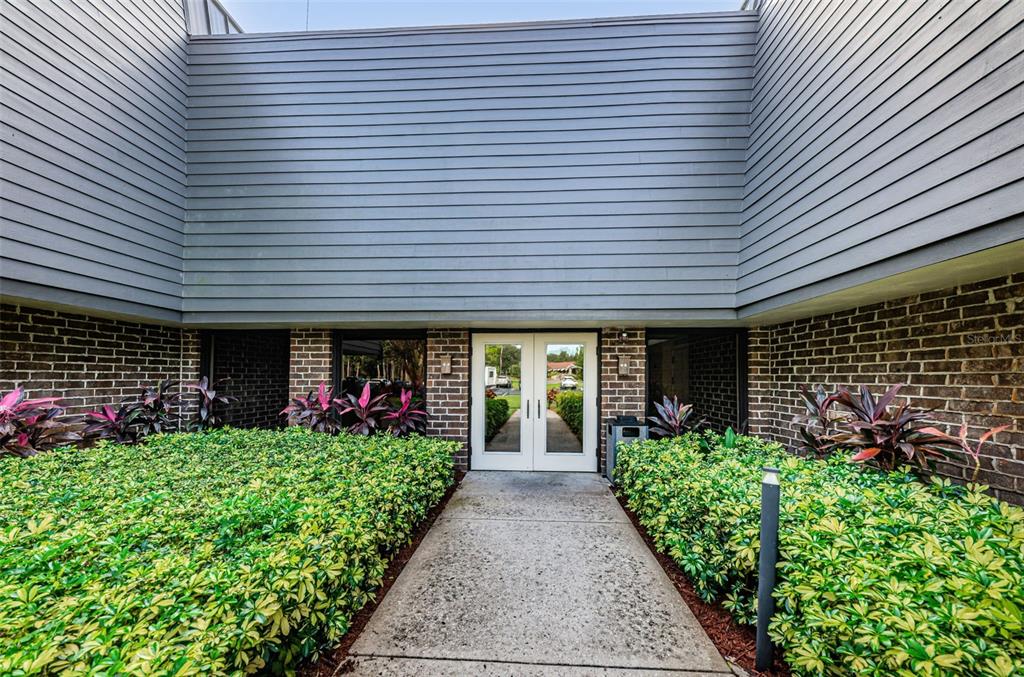 A concrete walkway leads to double glass doors centered between brick walls and gray siding — 36750 US HIGHWAY 19 N UNIT 5-104, PALM HARBOR, FL