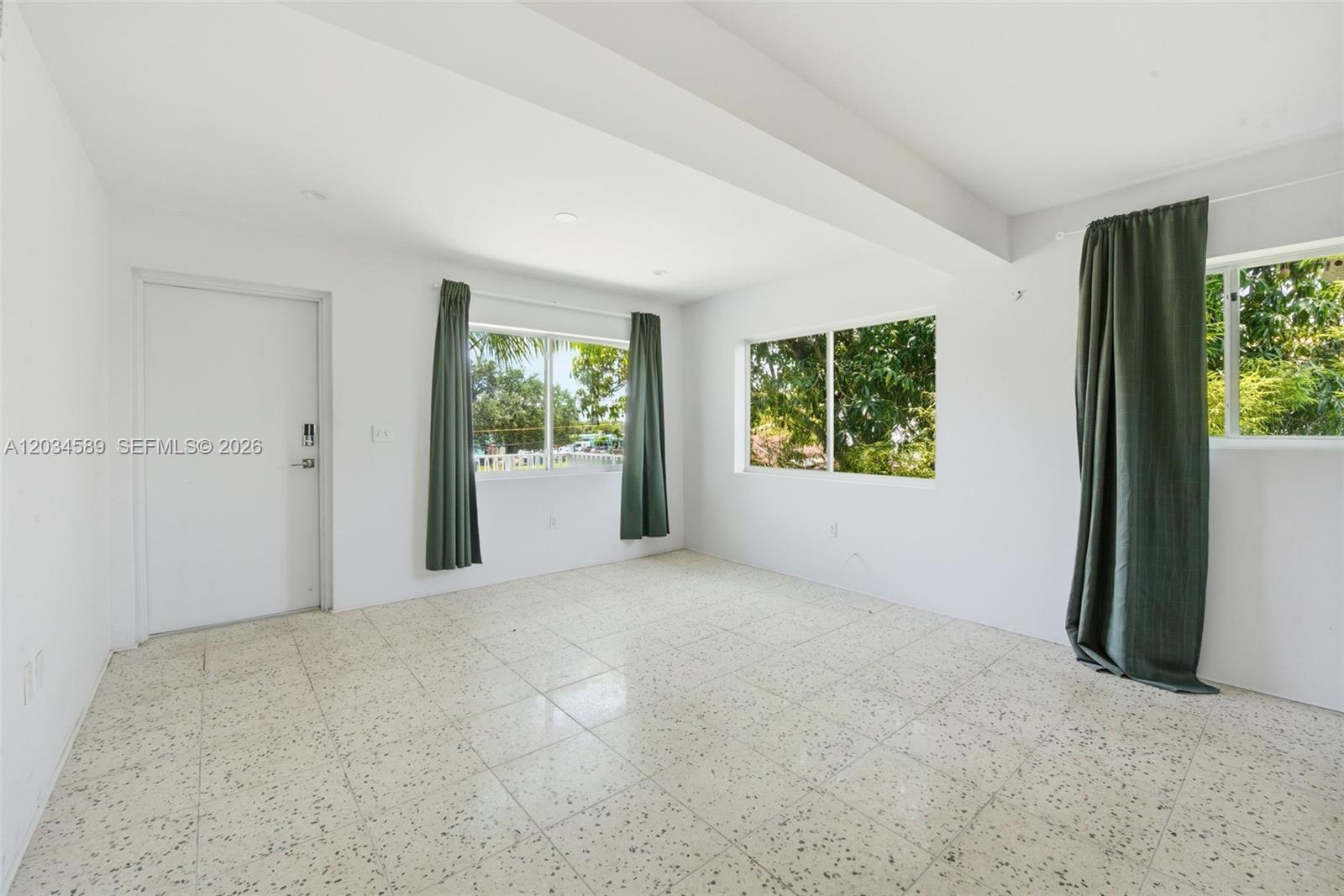 An empty room features white walls, speckled tile flooring, and windows with green curtains — 127 NW 74th St # 3, Miami FL 33150, MIAMI, FL