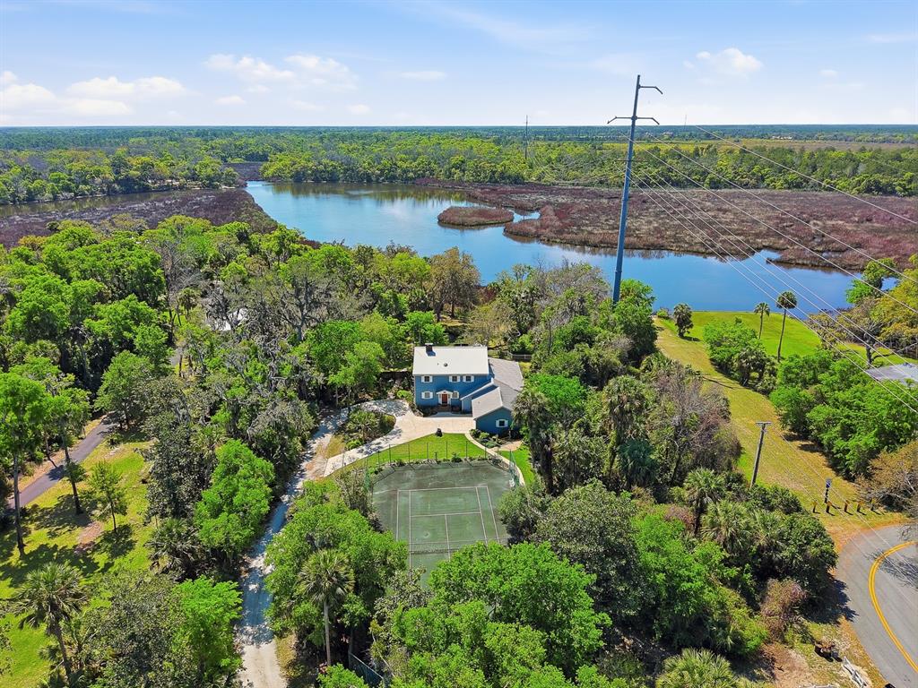 An aerial view shows a blue house with a tennis court near a lake — 6491 SPRUCE CREEK ROAD, PORT ORANGE, FL