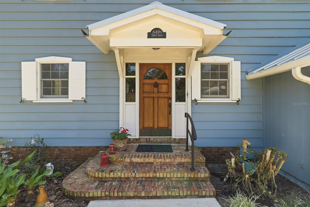 A blue-sided house features a brick porch, a wooden front door, and a white portico — 6491 SPRUCE CREEK ROAD, PORT ORANGE, FL