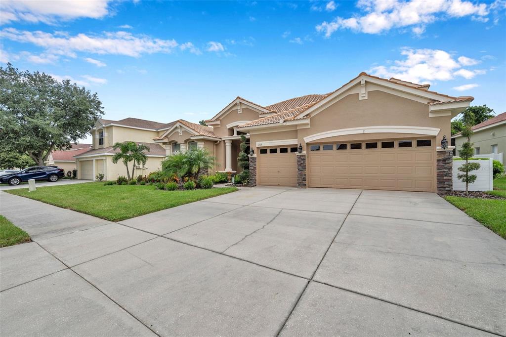 A tan single-story house features a three-car garage and a concrete driveway under a sky — 11519 BELLE HAVEN DRIVE, NEW PORT RICHEY, FL