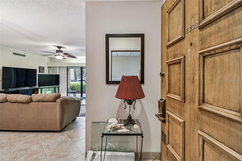 An entryway with a wooden door, a mirror, a lamp, and a view into a living room — 36750 US HIGHWAY 19 N UNIT 03106, PALM HARBOR, FL