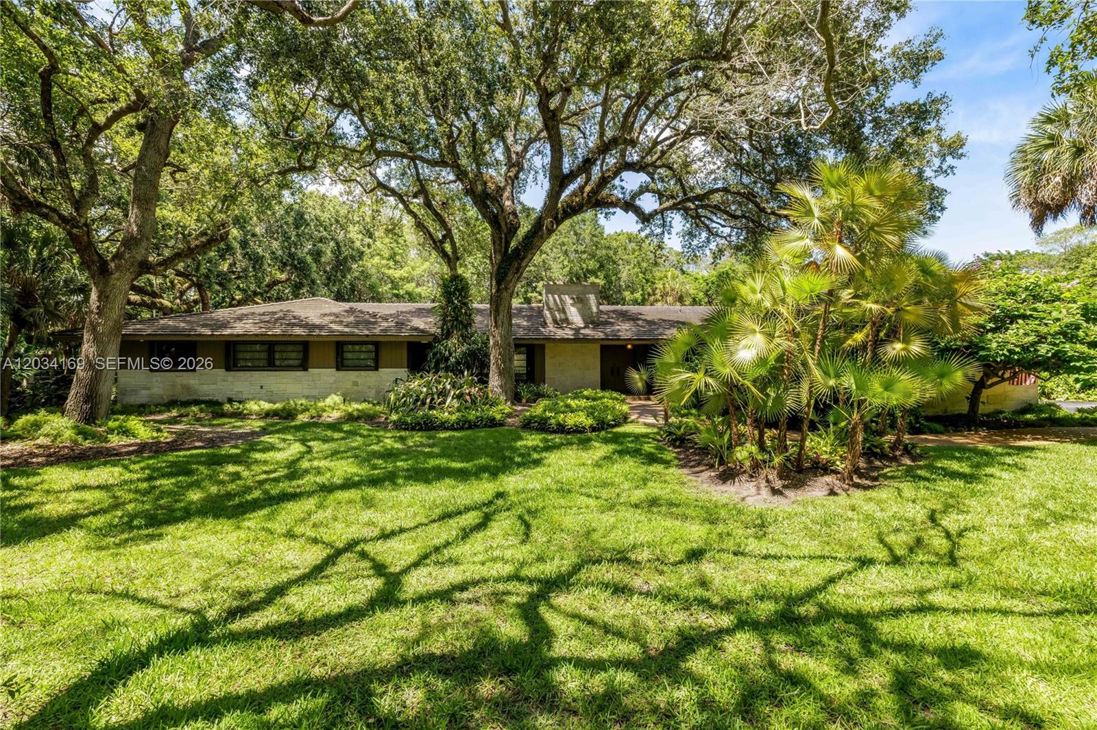 A single-story house with a stone facade sits behind a large lawn and mature trees — 9979 Fairchild Way, Coral Gables FL 33156, CORAL GABLES, FL