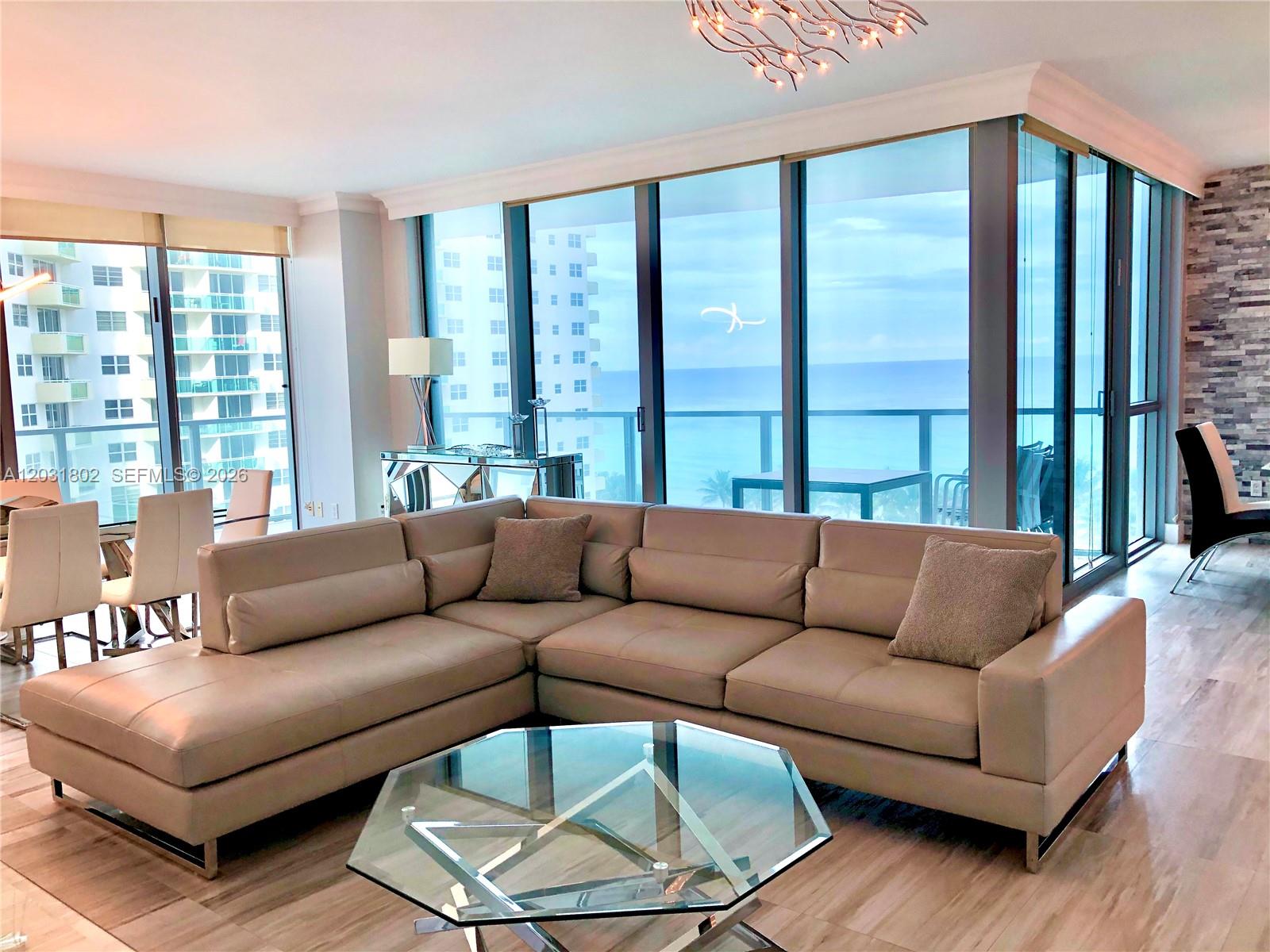 A modern living room features a beige sectional sofa, glass coffee table, and floor-to-ceiling windows — 3101 S Ocean Dr # 801, Hollywood FL 33019, HOLLYWOOD, FL