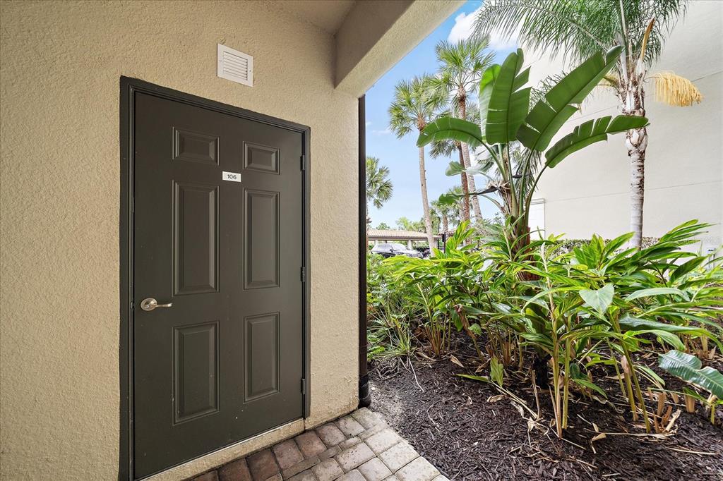 A dark brown door labeled 106 opens to a paved walkway beside lush tropical landscaping — 16814 VARDON TERRACE UNIT 106, BRADENTON, FL