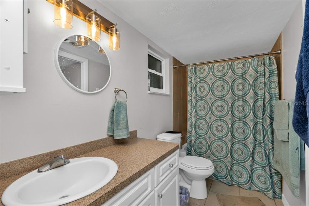 A bathroom features a white vanity with a tan countertop, a round mirror, and a shower — 3100 HARBOR BOULEVARD UNIT 108, PORT CHARLOTTE, FL