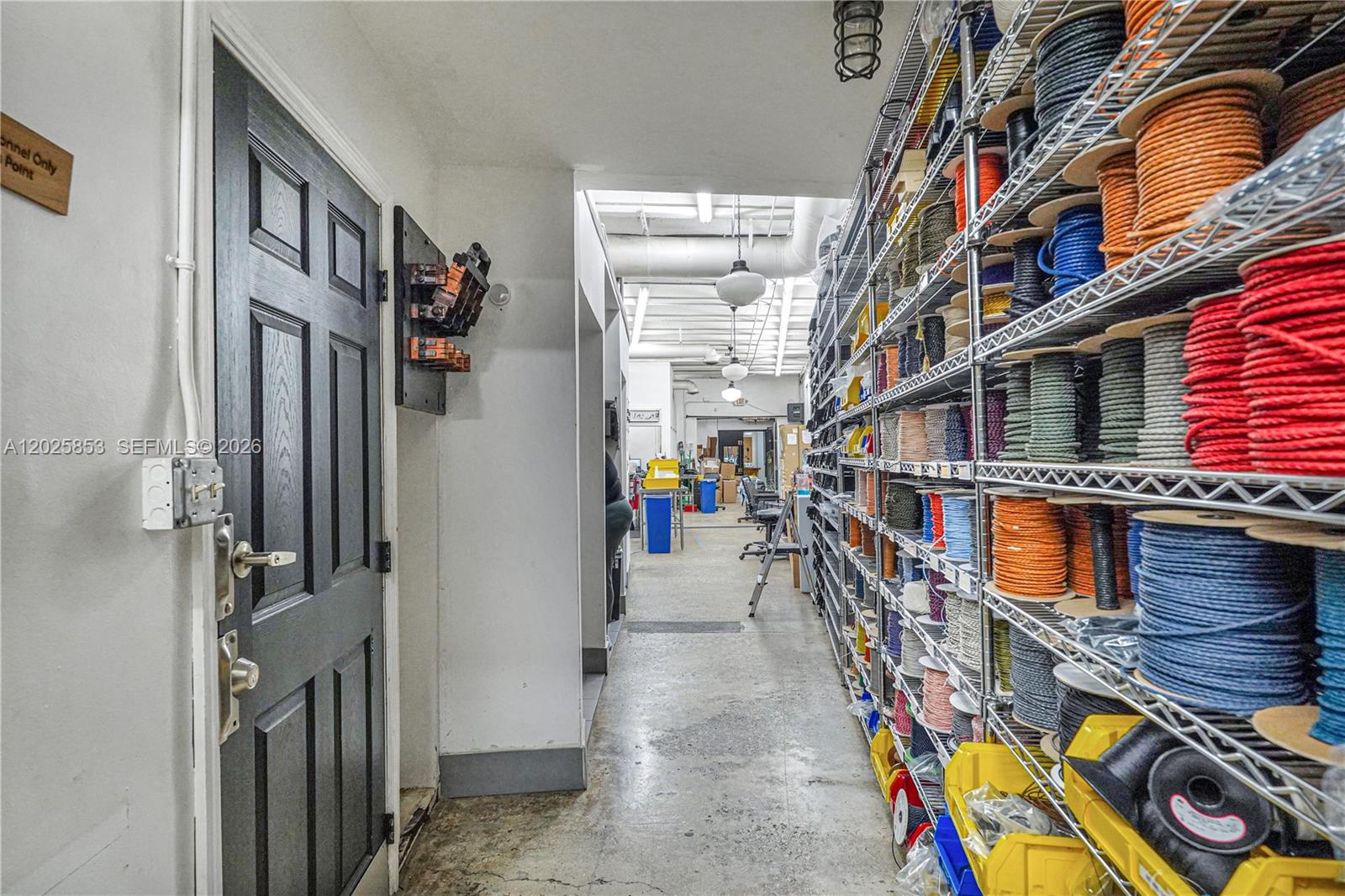 A hallway features a black door, concrete floors, and tall metal shelving filled with spools — MIAMI, FL