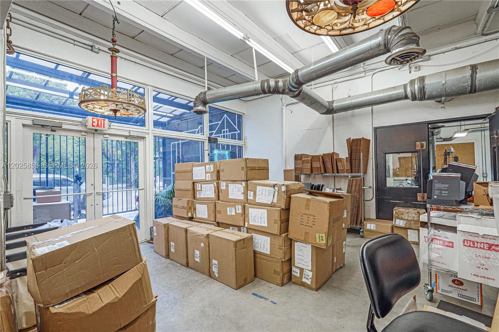 A commercial storage room features exposed ductwork, cardboard boxes, and glass doors with an exit sign — MIAMI, FL