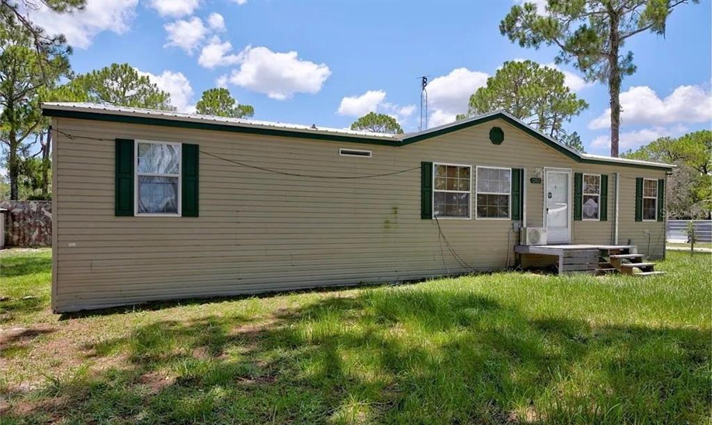 A tan manufactured home with green shutters sits on a grassy lot under a blue sky — 201 E MAXANNA, SEBRING, FL
