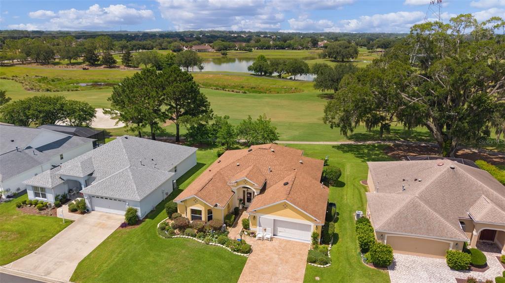 An aerial view shows three suburban houses with shingled roofs overlooking a golf course pond — 16840 SE 85TH SAPELO COURT, THE VILLAGES, FL
