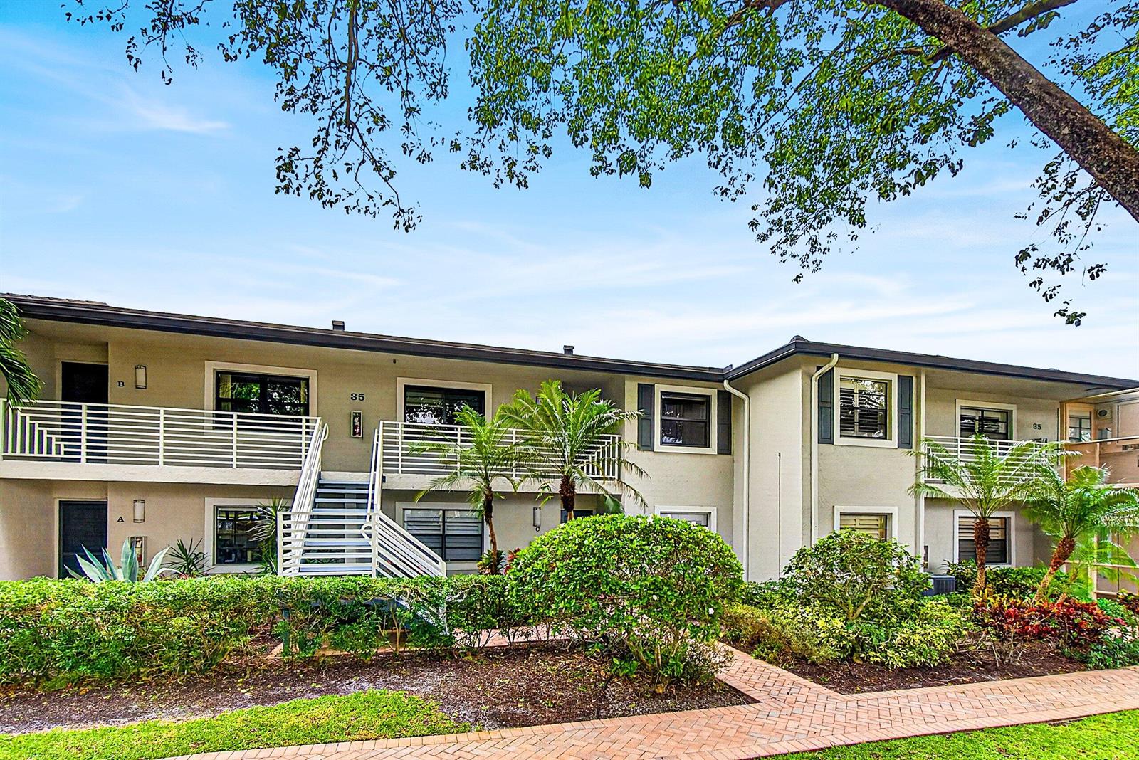 A two-story apartment building features beige stucco walls, white railings, and a brick walkway — 35 Southport Lane # D, Boynton Beach FL 33436, BOYNTON BEACH, FL