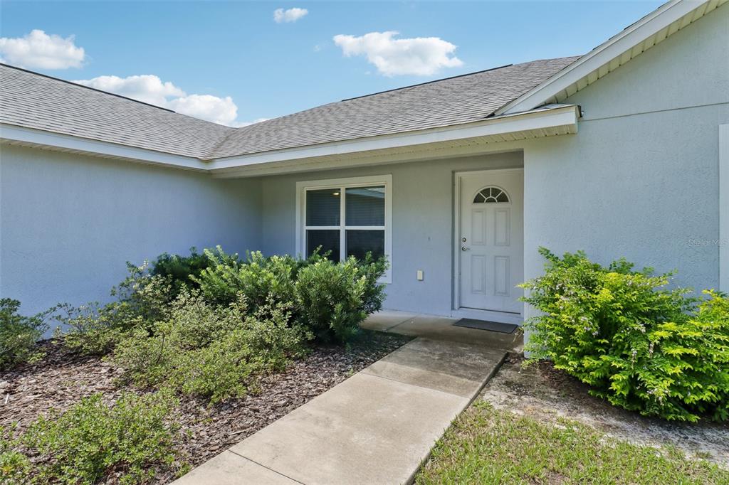A concrete walkway leads to the front entrance of a light-colored house with landscaping — 9170 SE 150TH STREET, SUMMERFIELD, FL