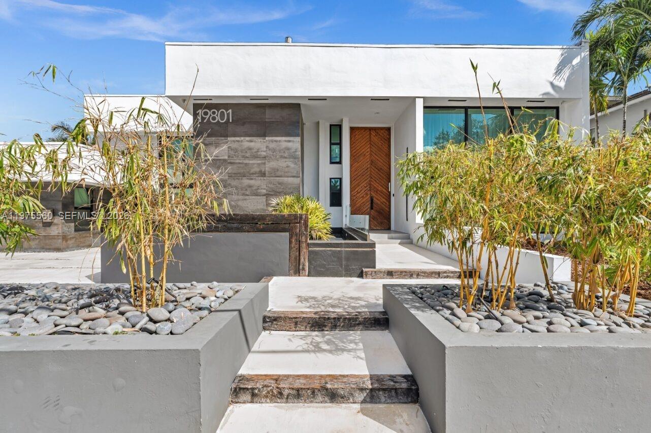 The exterior of a modern white house features a wooden front door and bamboo landscaping — 19801 NE 23rd Ave, Miami FL 33180, MIAMI, FL