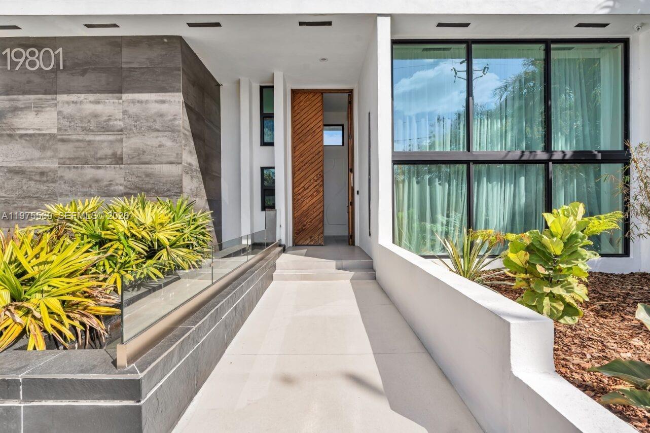 A modern home entrance features a diagonal wood door, glass railing, and large windows — 19801 NE 23rd Ave, Miami FL 33180, MIAMI, FL