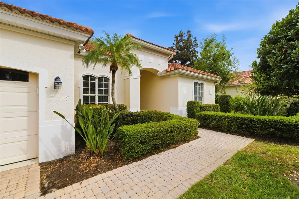 A beige stucco house features a tiled roof, a brick paver walkway, and manicured hedges — 7418 MONTE VERDE, SARASOTA, FL