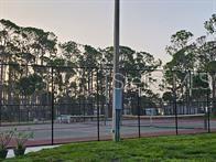 A fenced tennis court sits outdoors surrounded by tall pine trees under a clear sky — 5672 HOLIDAY PARK BOULEVARD, NORTH PORT, FL