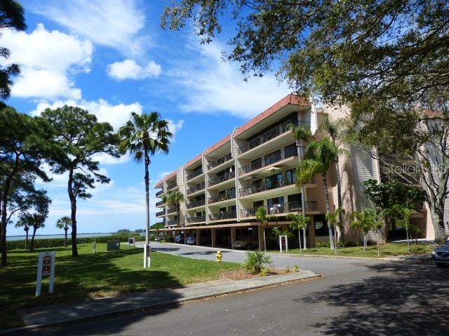 A multi-story apartment building stands near a grassy lawn and trees under a blue sky — 2700 BAYSHORE BOULEVARD UNIT 11307, DUNEDIN, FL
