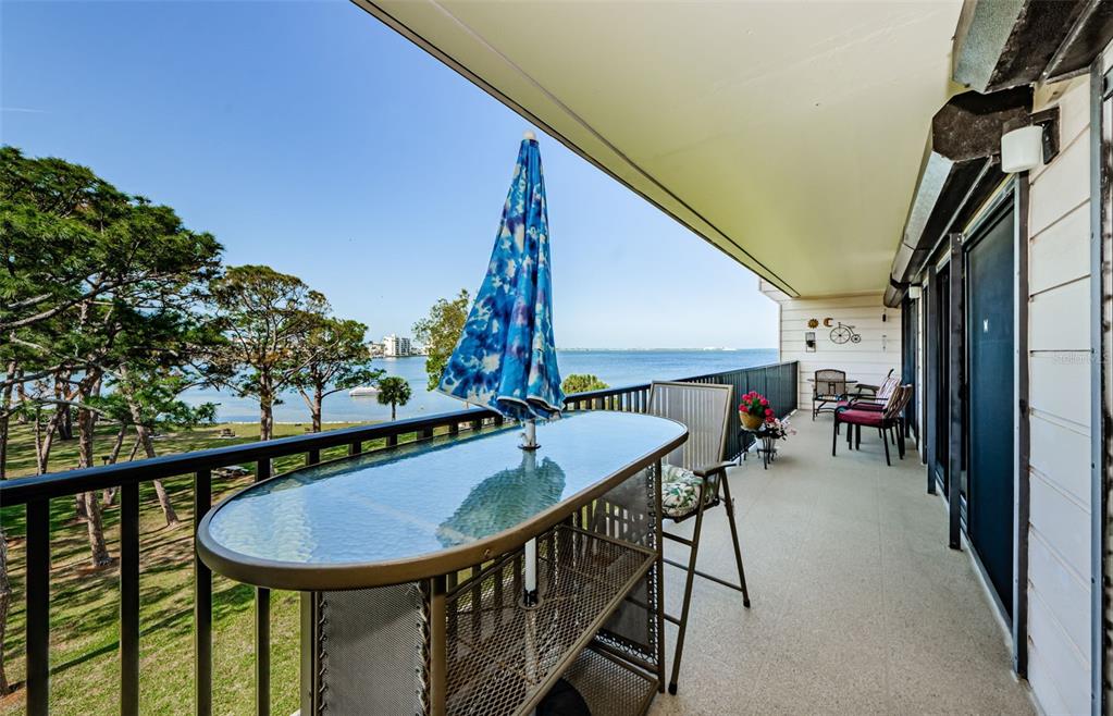 A balcony with a glass-topped table, umbrella, and chairs overlooking a park and water — 2700 BAYSHORE BOULEVARD UNIT 11307, DUNEDIN, FL