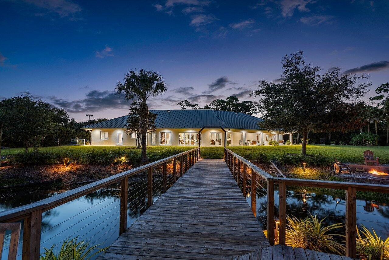 A wooden footbridge leads to a single-story house illuminated at dusk beside a pond — 6624 Dillman Road, FL 33413, FL