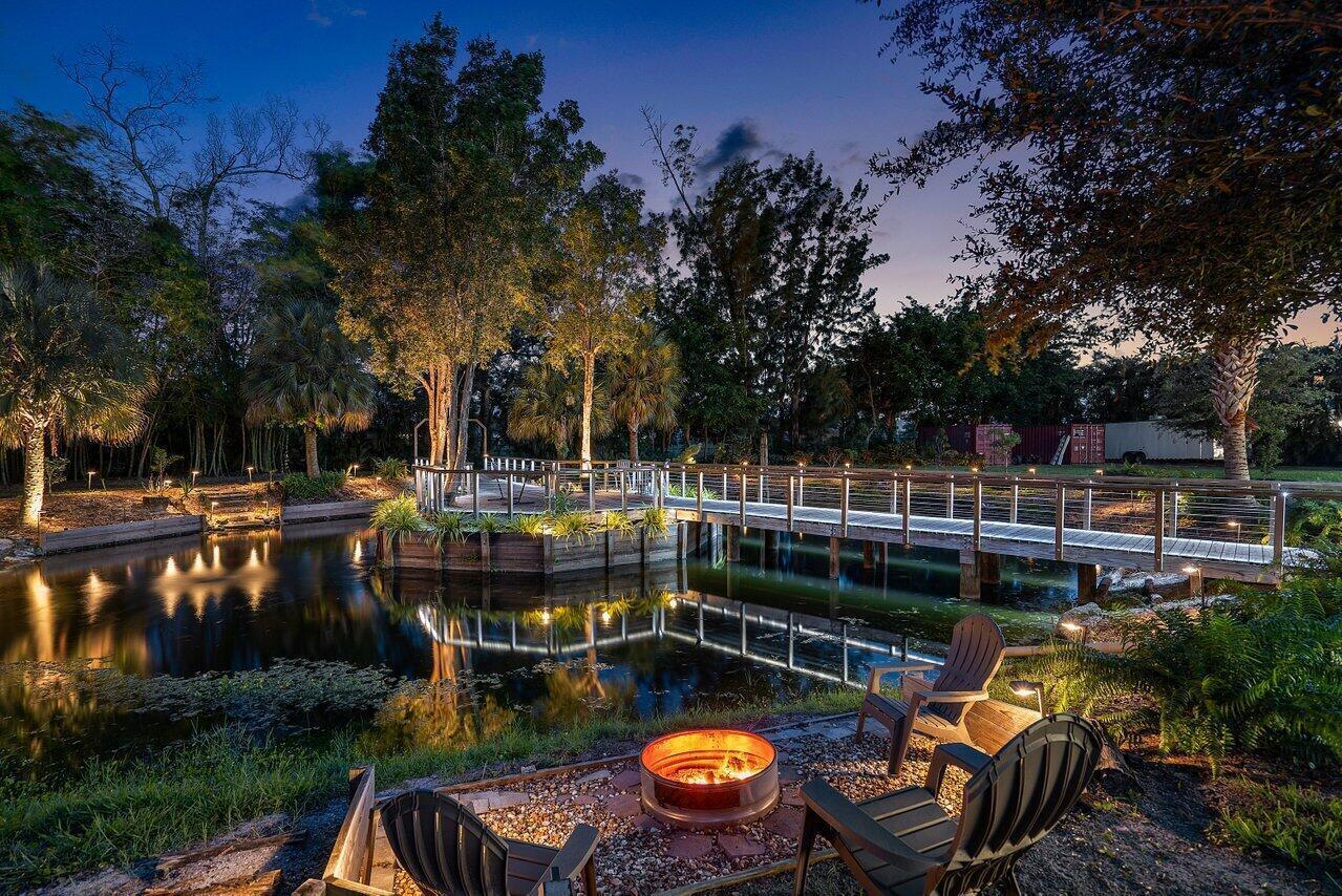A fire pit with chairs overlooks a pond and a wooden footbridge at dusk — 6624 Dillman Road, FL 33413, FL