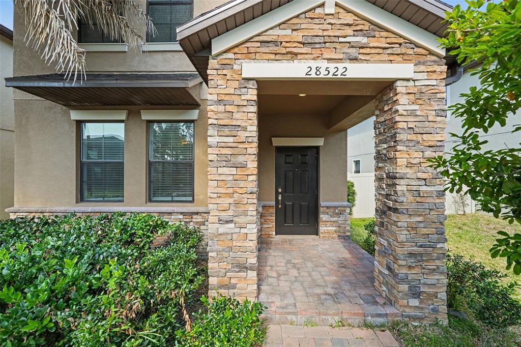 A stone-accented entryway features a dark front door, a brick paver porch, and surrounding landscaping — 28522 E PLEASANT BAY LOOP, WESLEY CHAPEL, FL