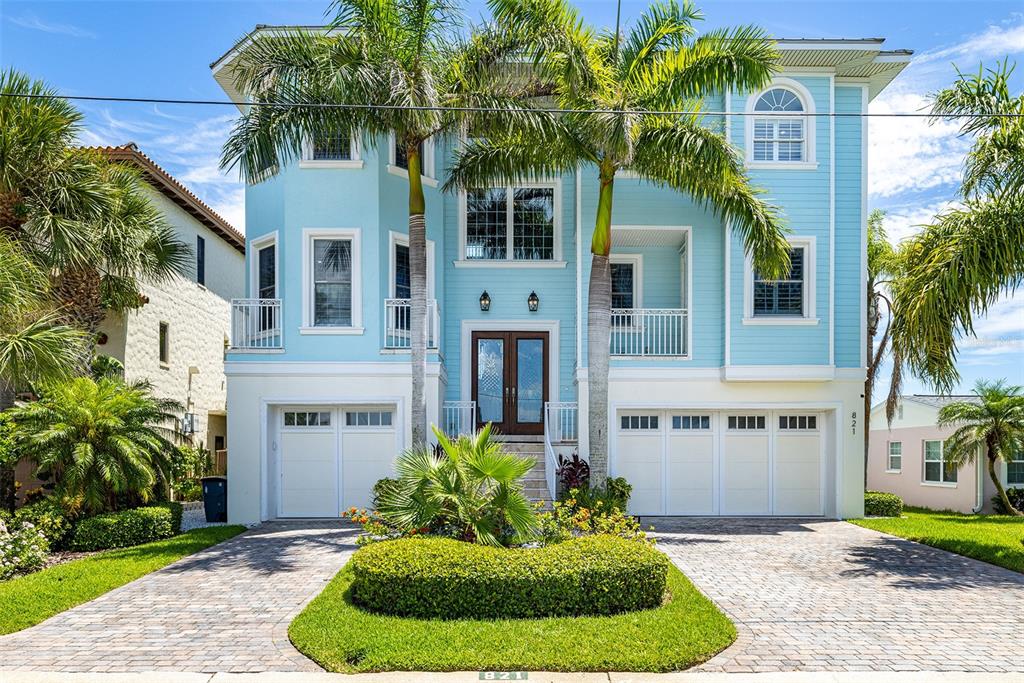 A three-story light blue house with a paved driveway, two garages, and palm trees — 821 BAY POINT DRIVE, MADEIRA BEACH, FL