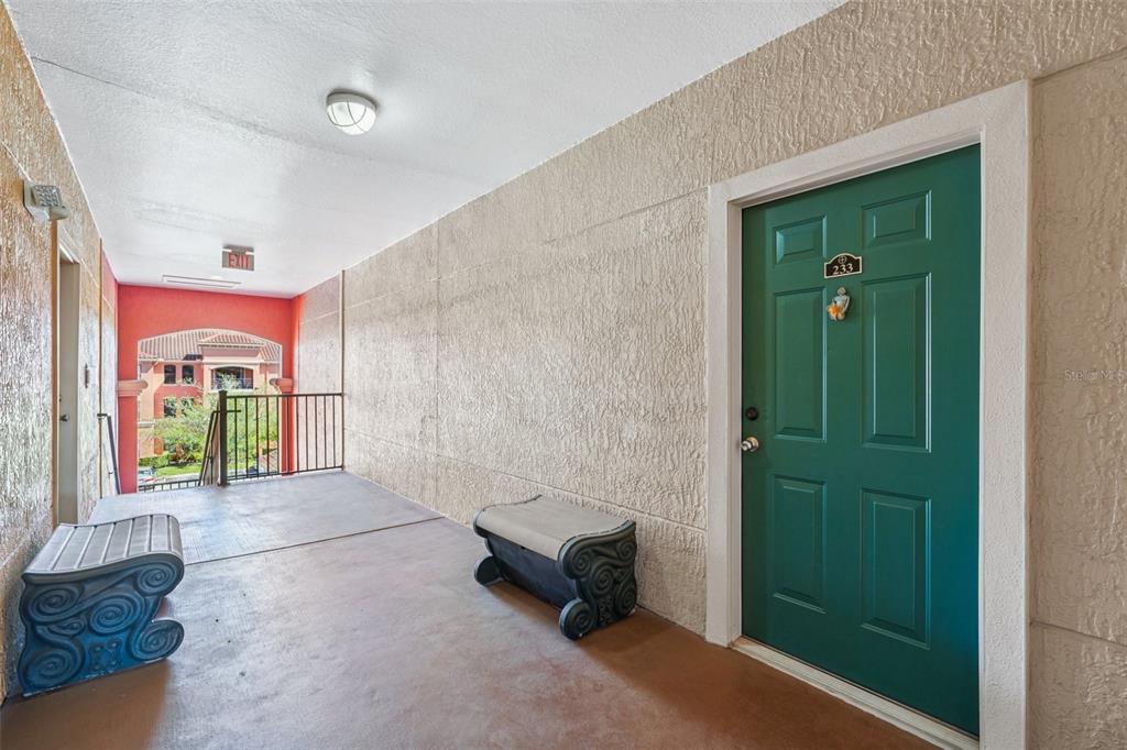An outdoor apartment hallway features a green door, two benches, and a view of buildings — 2713 VIA MURANO UNIT 233, CLEARWATER, FL