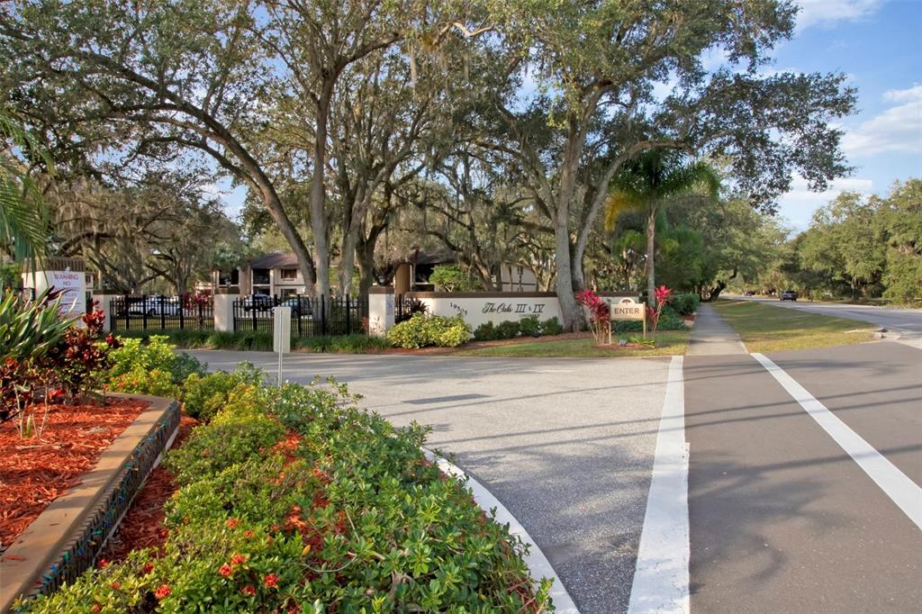 The entrance to a residential complex features a sign, landscaping, and large oak trees — 19505 QUESADA AVENUE UNIT J204, PORT CHARLOTTE, FL