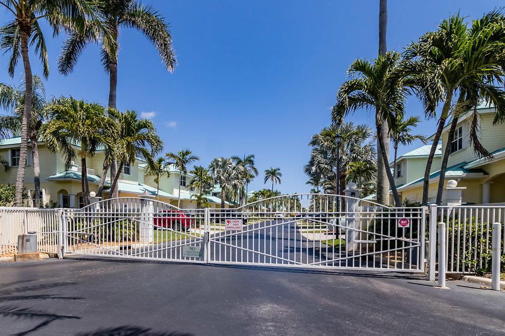 A white metal gate secures the entrance to a residential community with palm trees — 117 Barefoot Cove, Hypoluxo FL 33462, HYPOLUXO, FL