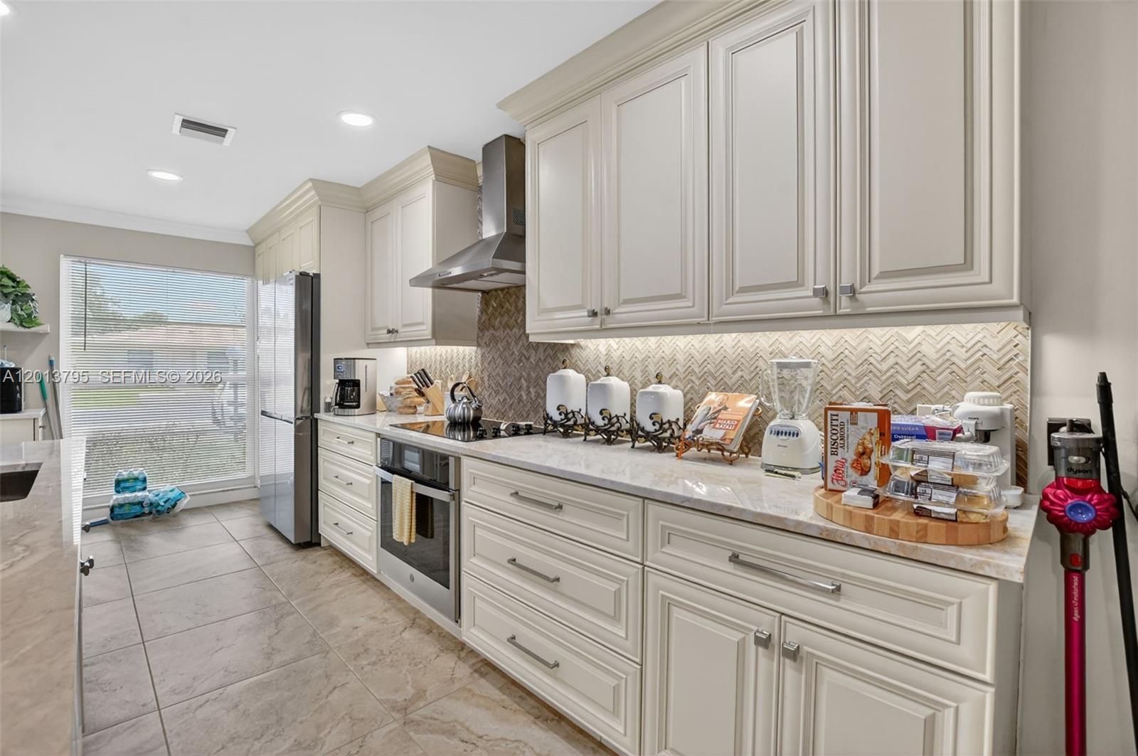 A kitchen features off-white cabinetry, stainless steel appliances, a herringbone backsplash, and tile flooring — 8749 Belle Aire Dr, Boca Raton FL 33433, BOCA RATON, FL