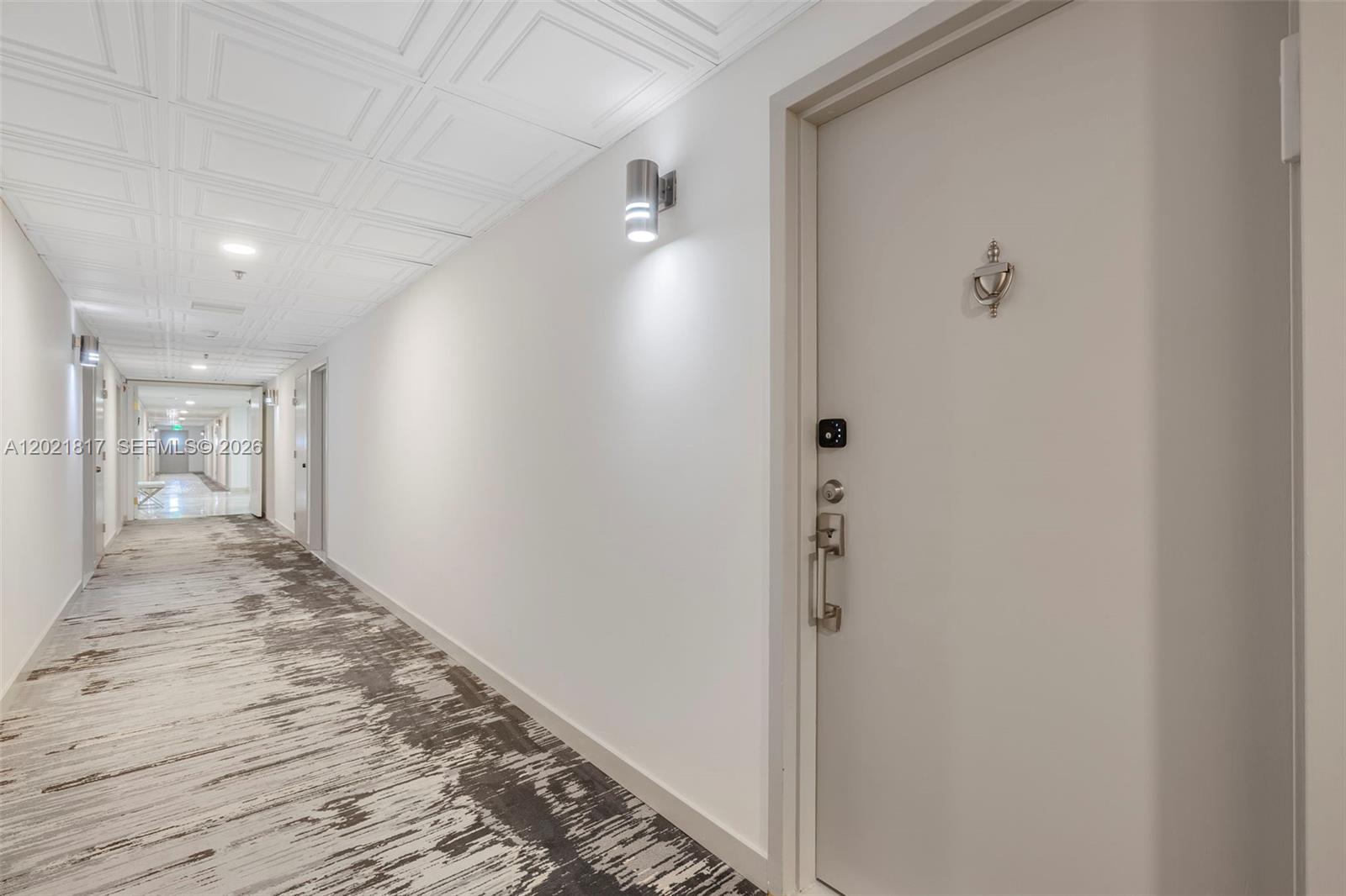 A long, brightly lit hallway features white walls, a patterned carpet, and a closed door — 9511 Collins Ave # 1202, Surfside FL 33154, SURFSIDE, FL