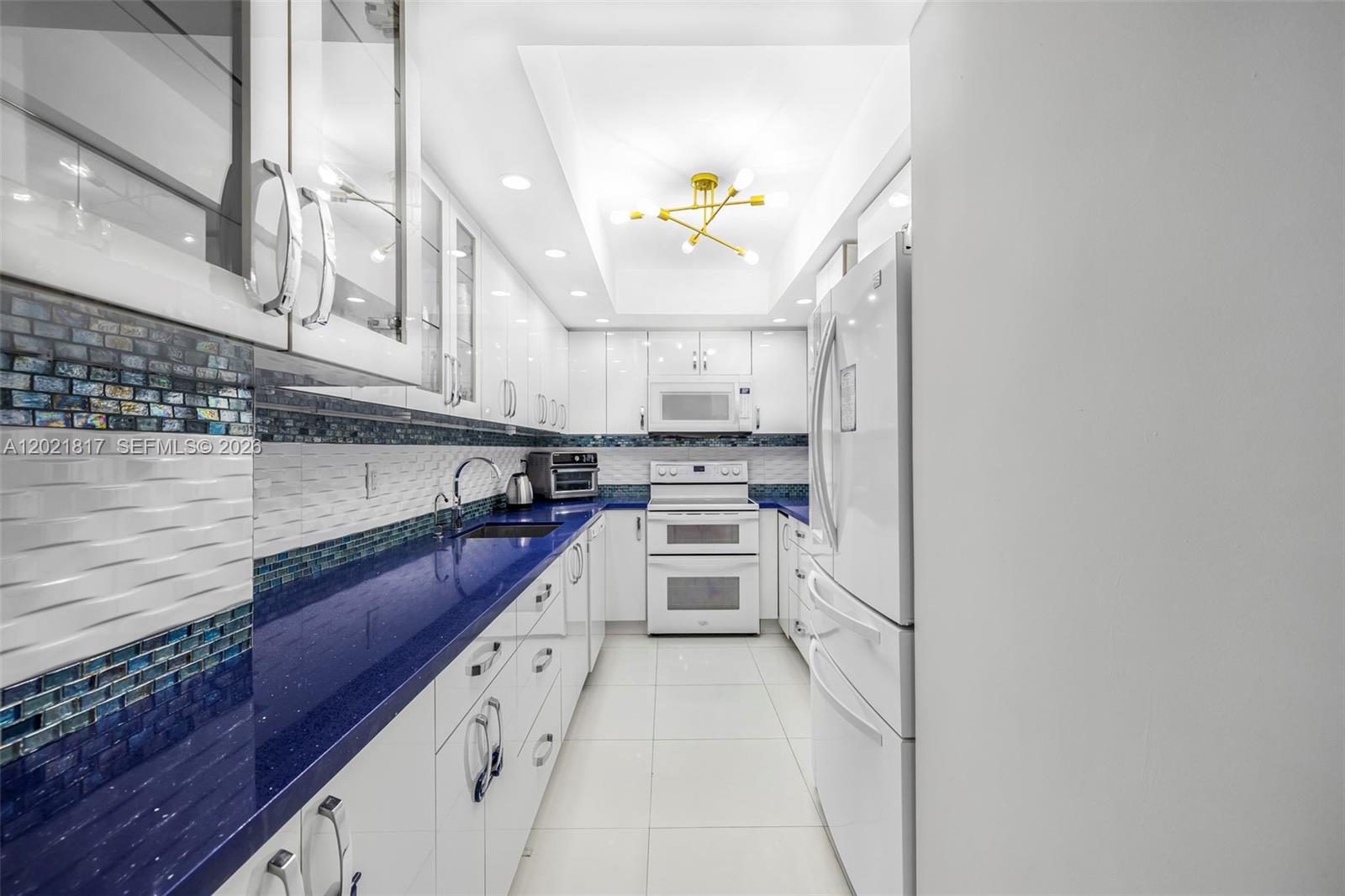 A galley kitchen features white cabinetry, blue countertops, a tiled backsplash, and white appliances — 9511 Collins Ave # 1202, Surfside FL 33154, SURFSIDE, FL