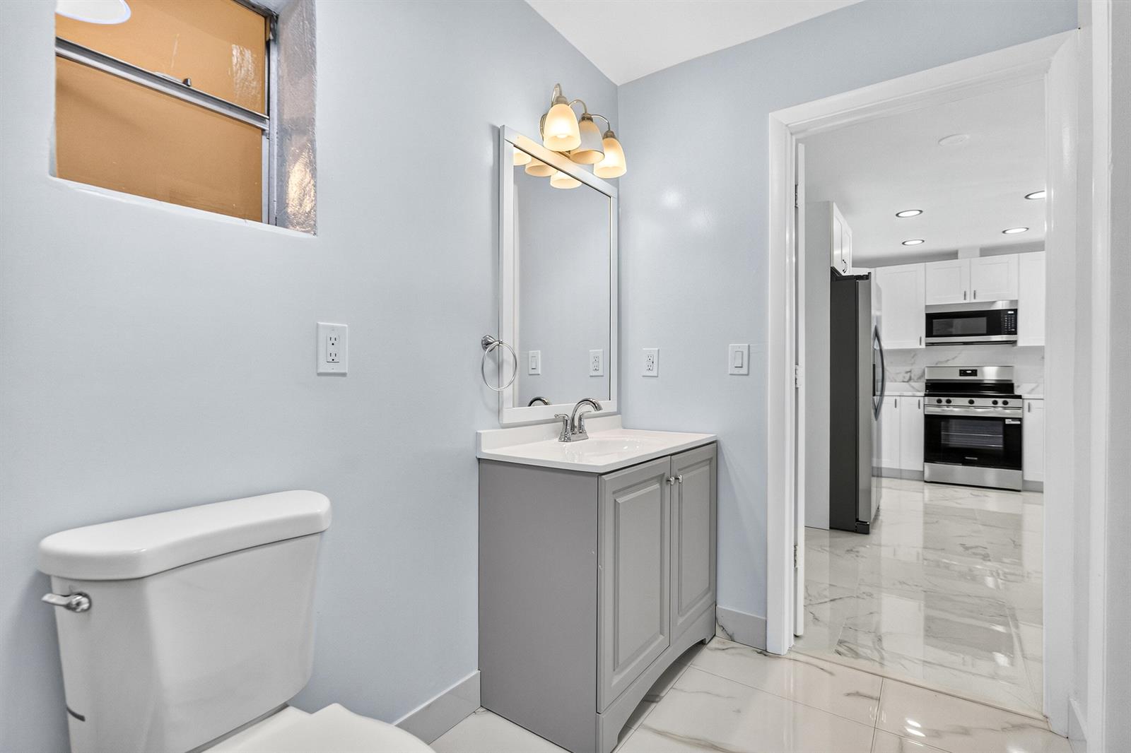 A bathroom with a gray vanity, white countertop, and toilet, looking into a white kitchen — 1531 NE 33rd Street, Pompano Beach FL 33064, POMPANO BEACH, FL