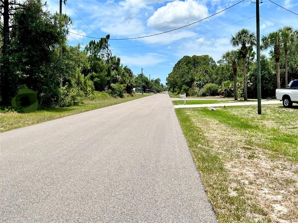A paved road stretches into the distance, flanked by trees, palm trees, and a grassy shoulder — 324 CORY STREET, PORT CHARLOTTE, FL