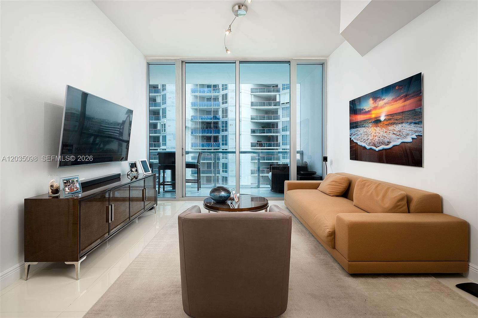 A modern living room features a tan sofa, a television, and a balcony with city views — 6799 Collins Ave # 510, Miami Beach FL 33141, MIAMI BEACH, FL