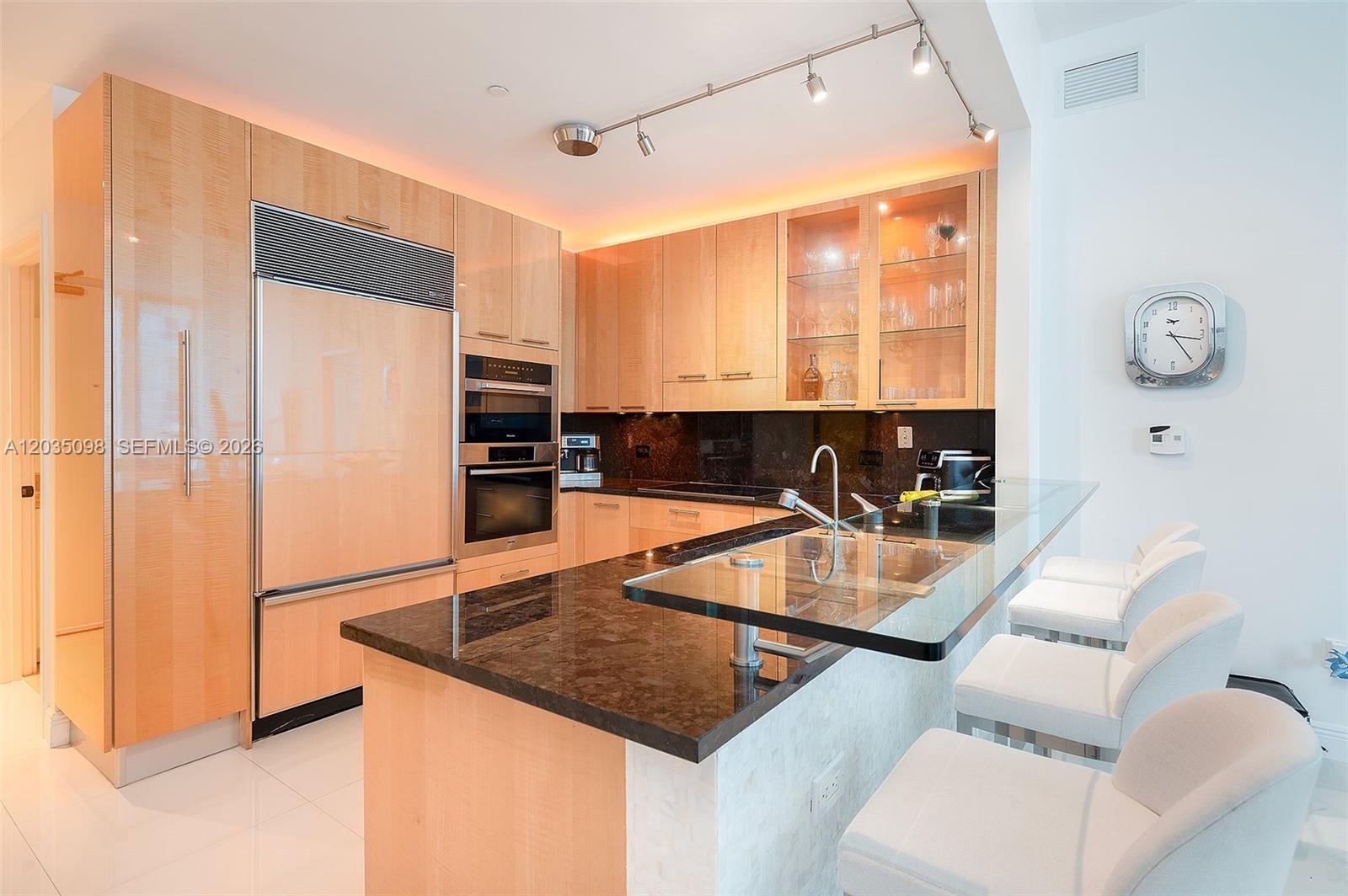A modern kitchen features light wood cabinetry, dark granite countertops, and a glass-topped breakfast bar — 6799 Collins Ave # 510, Miami Beach FL 33141, MIAMI BEACH, FL